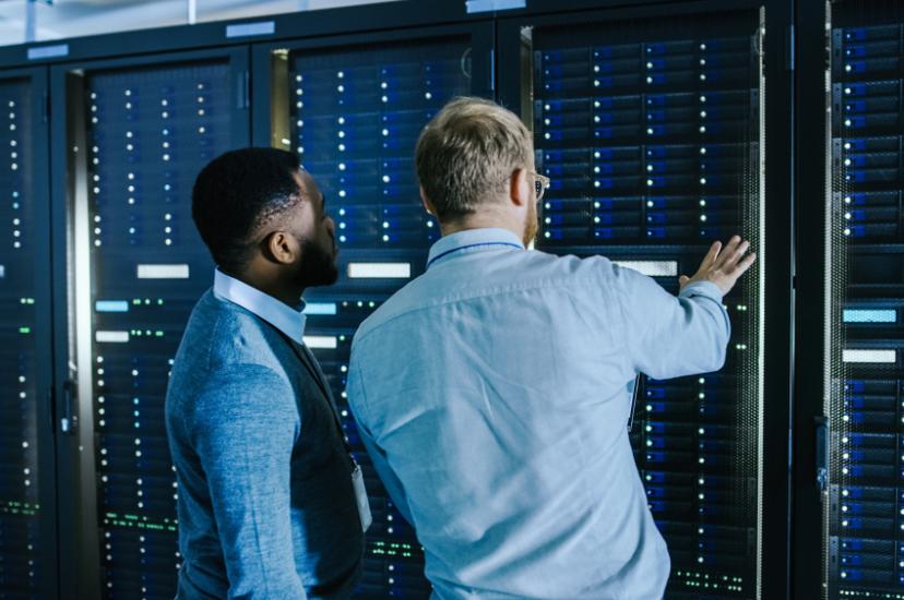 Bearded IT Technician in Glasses with a Laptop Computer and Male Engineer Colleague are Talking in Data Centre while Working Next to Server Racks. Running Diagnostics or Doing Maintenance Work