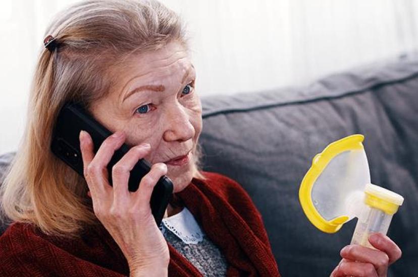 A concerned older woman holding an inhaler while talking on a mobile phone, seated on a sofa