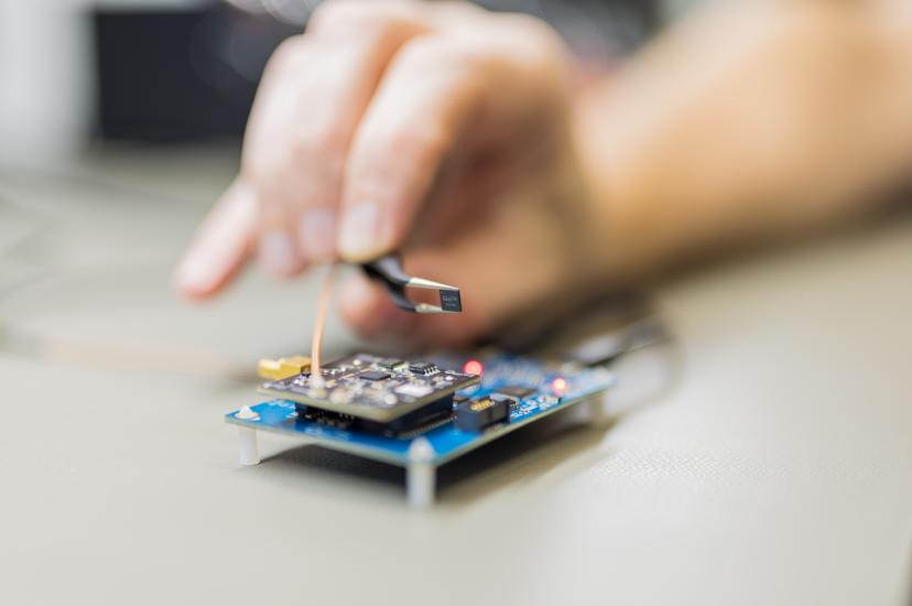 A pair of black forceps holds a tiny, square Qualinx QLX3Gx GNSS receiver chip over a blue electronic circuit board