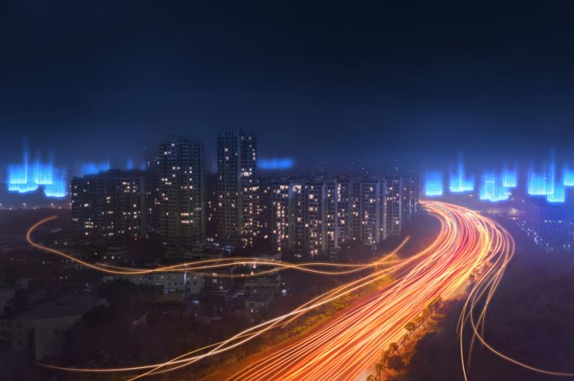 A long-exposure night shot of a city skyline featuring vibrant orange light trails from highway traffic and ethereal blue digital light effects floating above the buildings.