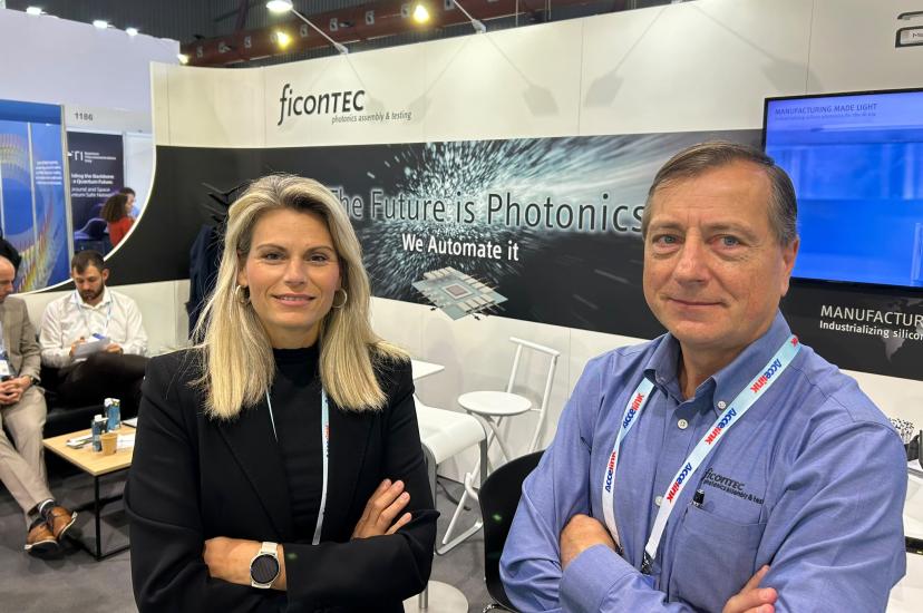 A blonde white woman in a dark jacket and a white man in a clean shirt stand in a conference booth