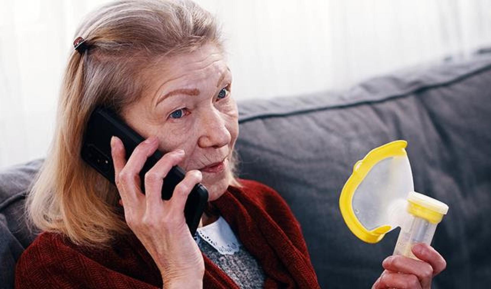 A concerned older woman holding an inhaler while talking on a mobile phone, seated on a sofa