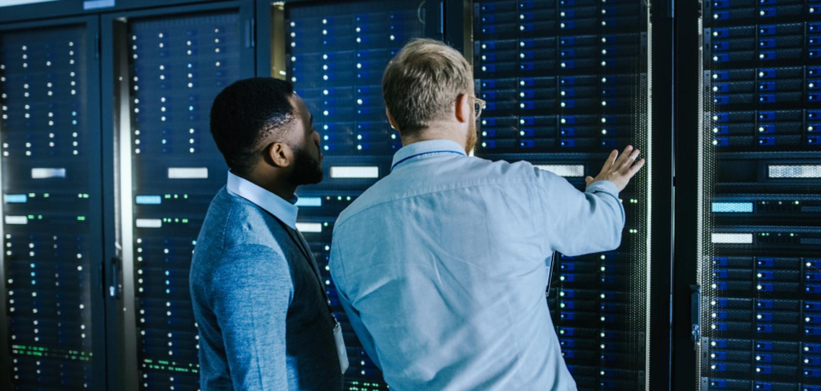 Bearded IT Technician in Glasses with a Laptop Computer and Male Engineer Colleague are Talking in Data Centre while Working Next to Server Racks. Running Diagnostics or Doing Maintenance Work