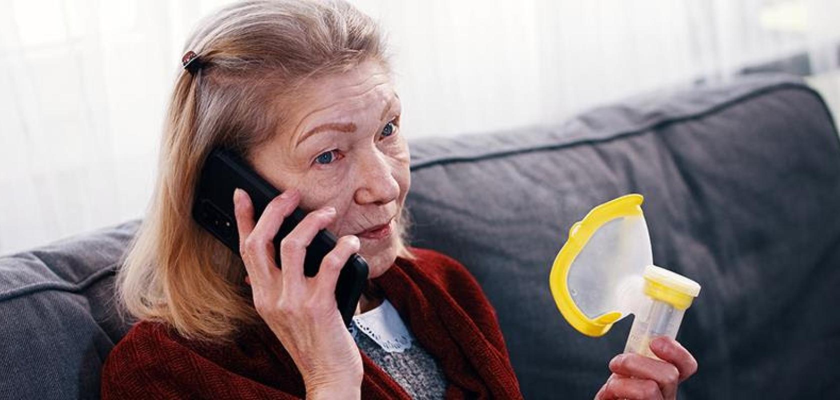 A concerned older woman holding an inhaler while talking on a mobile phone, seated on a sofa