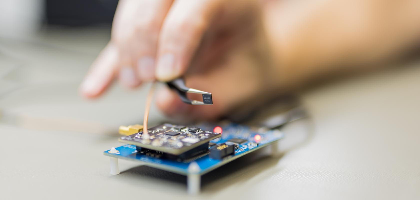 A pair of black forceps holds a tiny, square Qualinx QLX3Gx GNSS receiver chip over a blue electronic circuit board