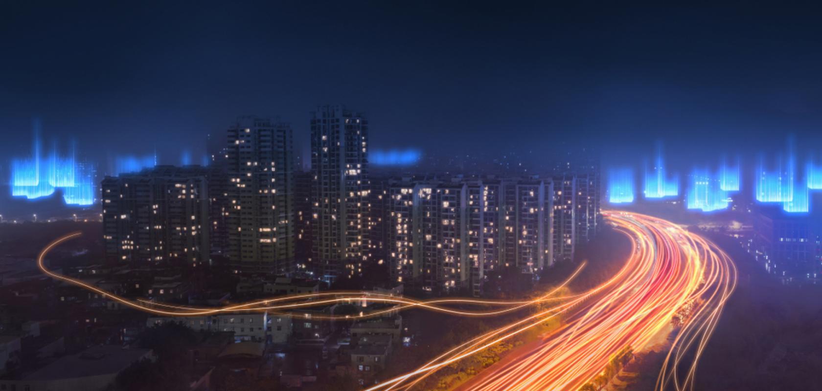 A long-exposure night shot of a city skyline featuring vibrant orange light trails from highway traffic and ethereal blue digital light effects floating above the buildings.