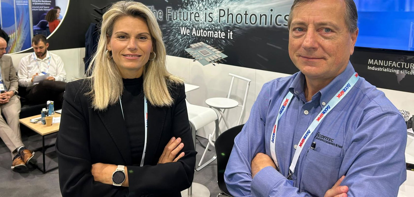 A blonde white woman in a dark jacket and a white man in a clean shirt stand in a conference booth