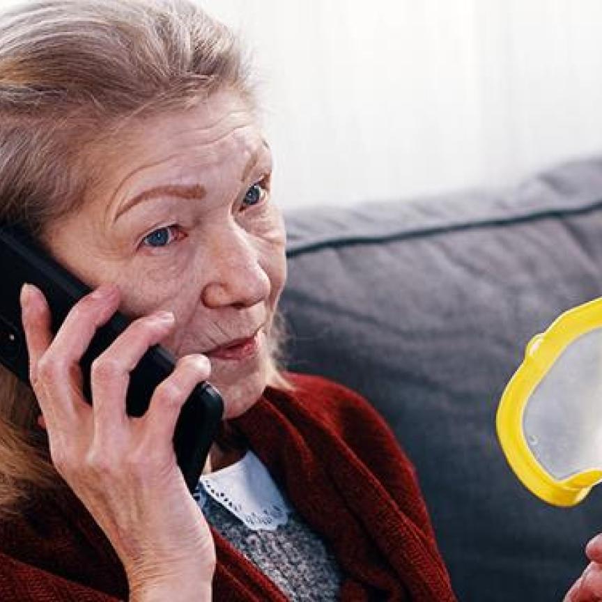 A concerned older woman holding an inhaler while talking on a mobile phone, seated on a sofa