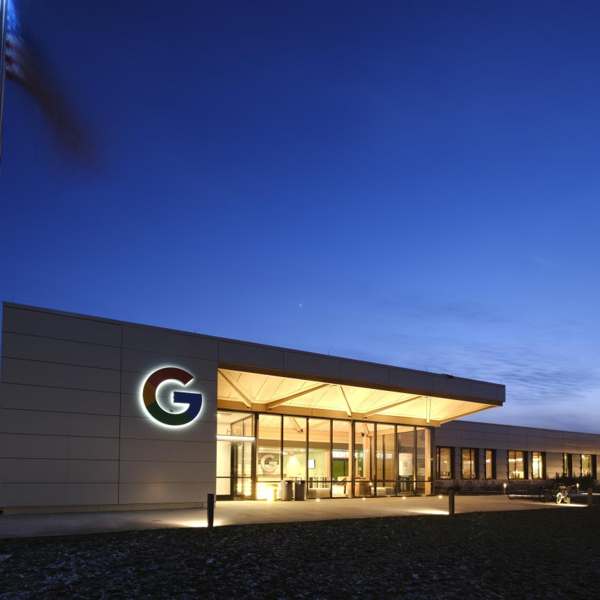 Exterior view of the Google New Albany data centre hub at twilight, featuring the illuminated Google logo on a modern white building facade with an American flag in the foreground